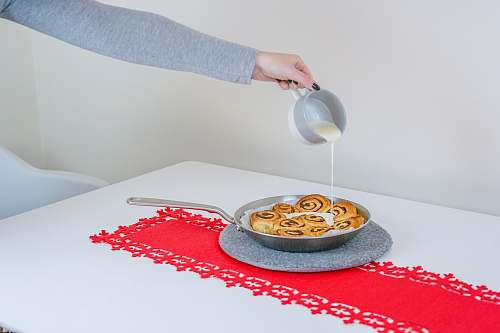 food person in gray long-sleeved shirt holding cup and pouring white liquid on plate with brown pastry cinnamon buns